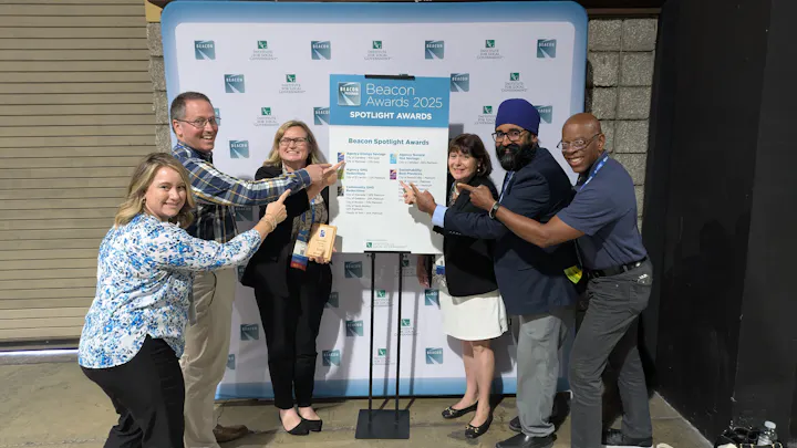 A group of people at an event celebrate the Beacon Awards 2025, pointing at a display of award categories and recipients.