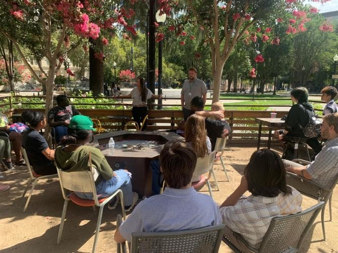 A group of people are sitting outdoors under trees, listening to two speakers standing near a wooden bench.