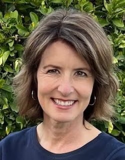 A woman with short brown hair smiling in front of leafy greenery.