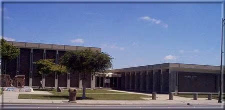 A modern building with pillars and a small courtyard, accompanied by trees and a clear blue sky.