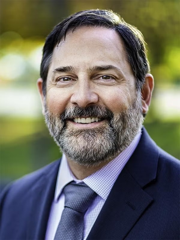 A smiling man in a suit with a beard, with a blurred outdoor background.