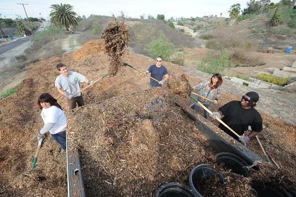 A group of people using shovels to move a large pile of mulch outdoors.