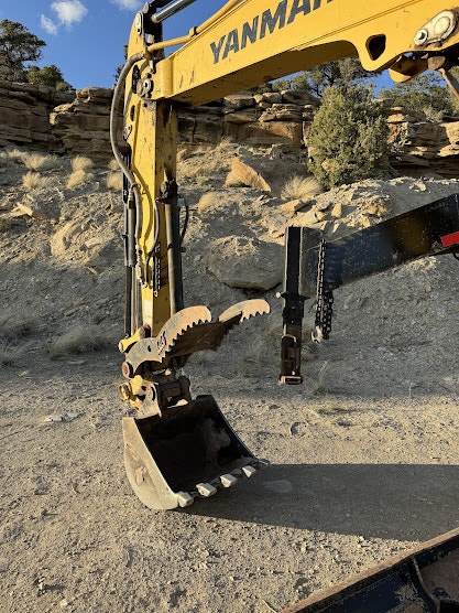 Excavator arm with a bucket and hydraulic thumb attachment, rocky terrain in the background.