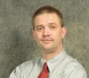 A man with light stubble wearing a button-up shirt and a red tie, in front of a mottled background.