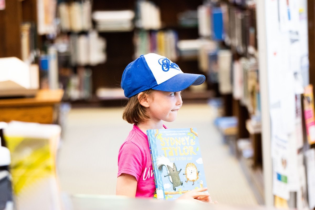 May contain: baseball cap, cap, clothing, hat, book, publication, indoors, library, person, and reading