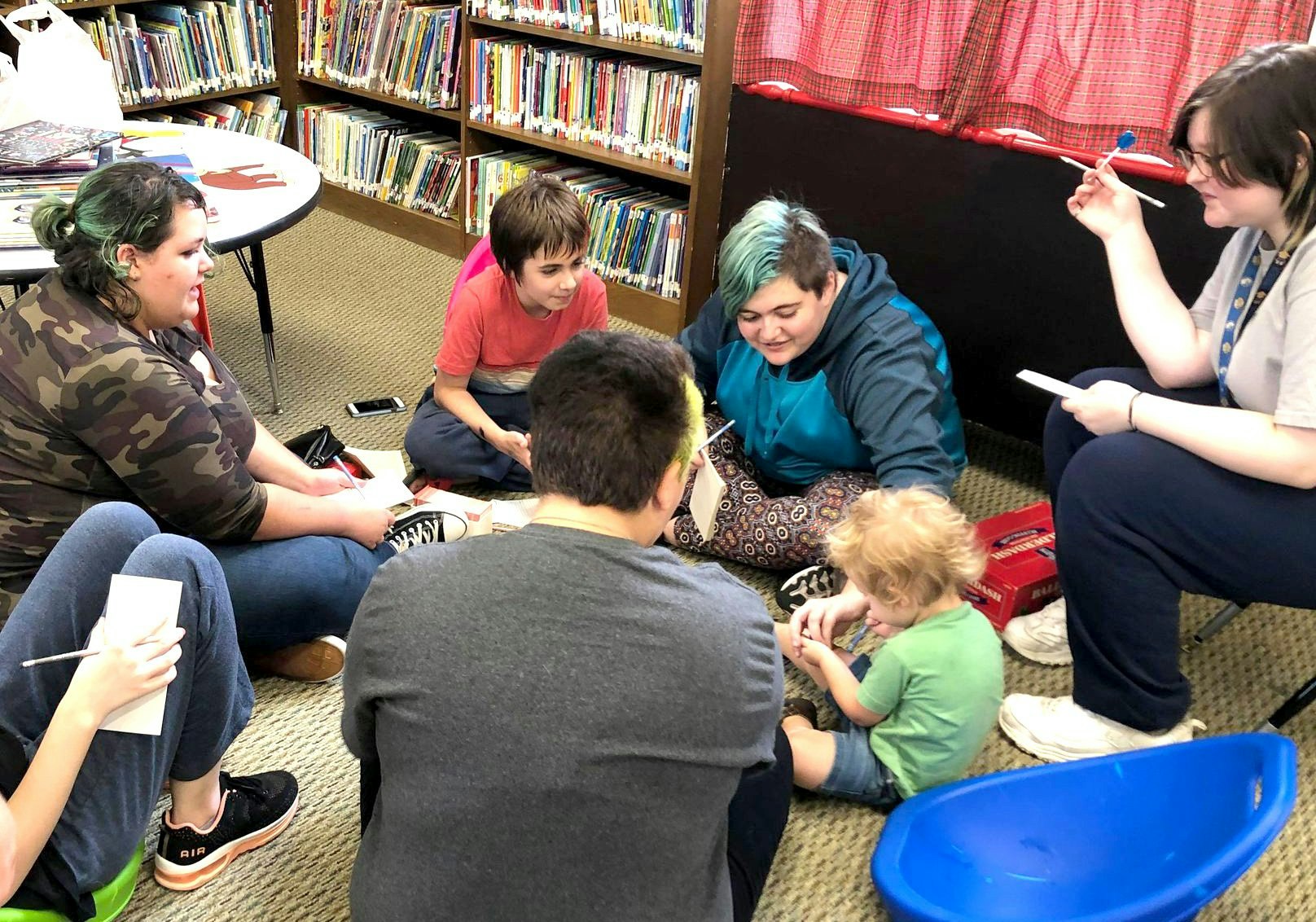 A group of people of various ages sitting on a carpet, engaging in what appears to be an activity or discussion, surrounded by bookshelves.