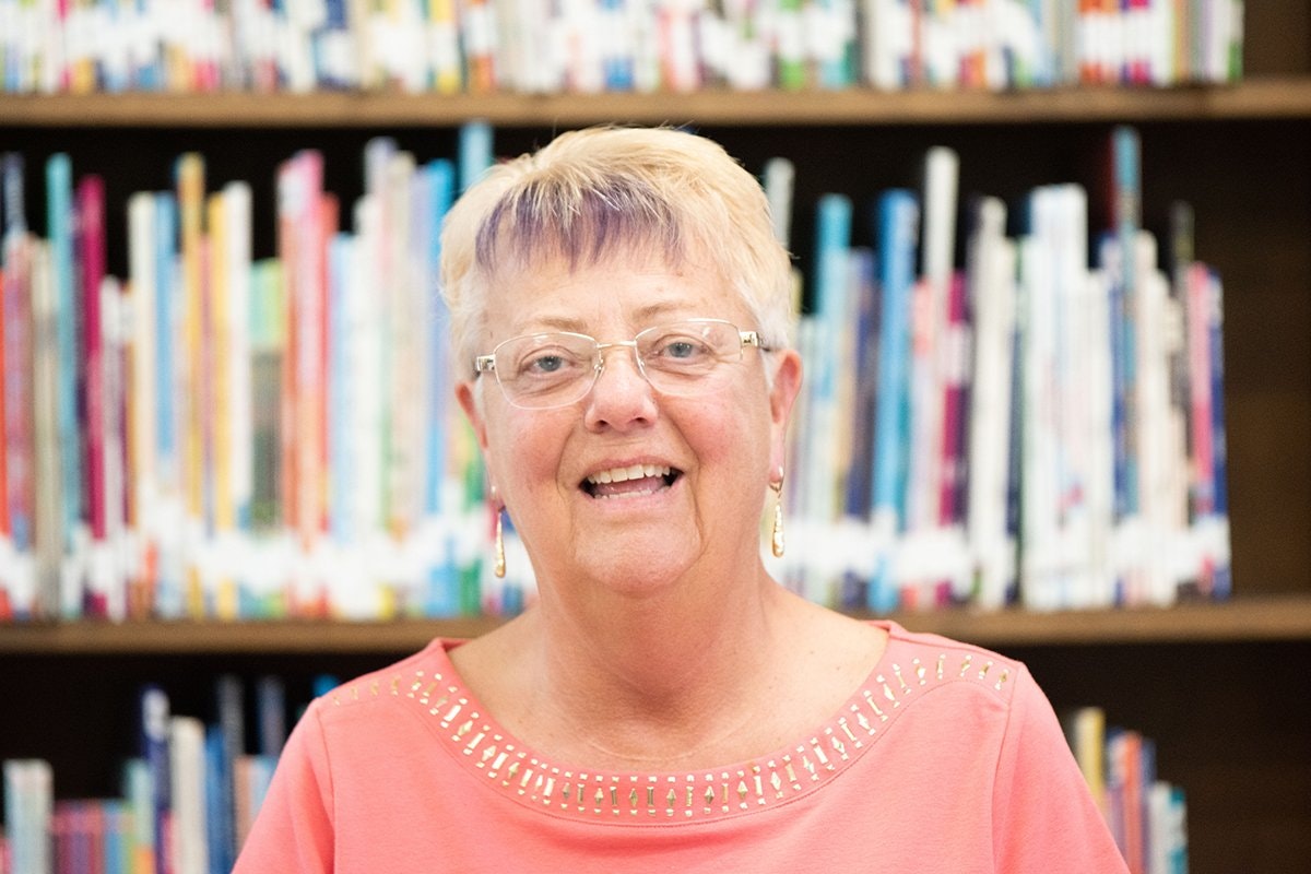 A smiling older woman with light blonde hair and glasses poses in front of a colorful bookshelf filled with books.