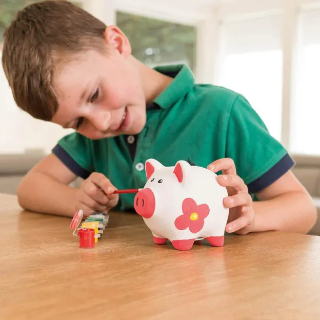 A young boy is painting a white piggy bank with colorful paints, focused on his creative activity at a table.