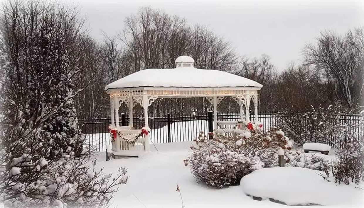 A snowy gazebo decorated with holiday garlands and lights, surrounded by snow-covered trees and bushes.