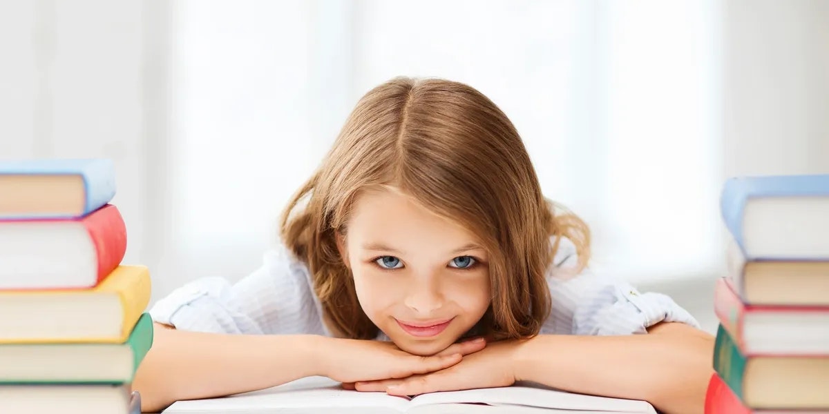A girl with brown hair is smiling, resting her head on a book, surrounded by stacked books.