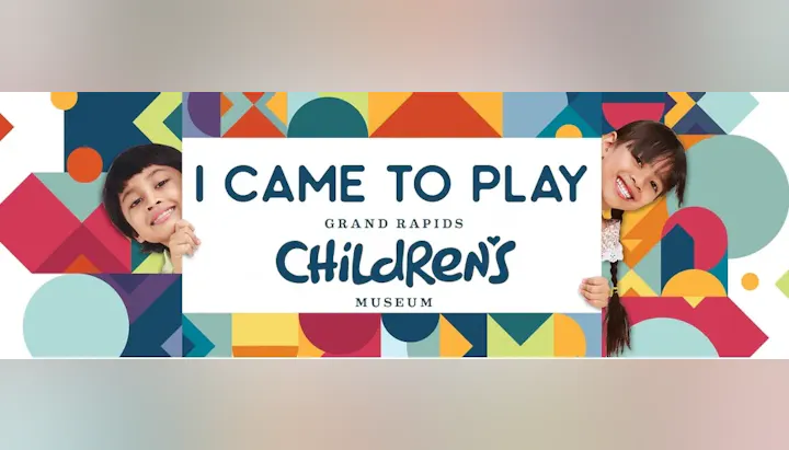 Two children peeking behind a sign: "I Came to Play, Grand Rapids Children's Museum," with colorful geometric patterns.