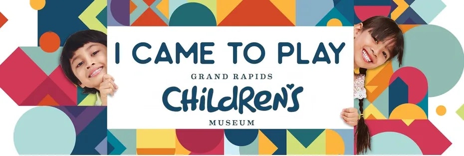 Two children peeking behind a sign: "I Came to Play, Grand Rapids Children's Museum," with colorful geometric patterns.