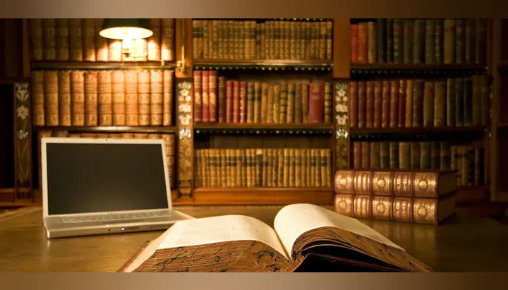 An open book on a desk, a laptop, and stacks of vintage books under warm light in a library setting.