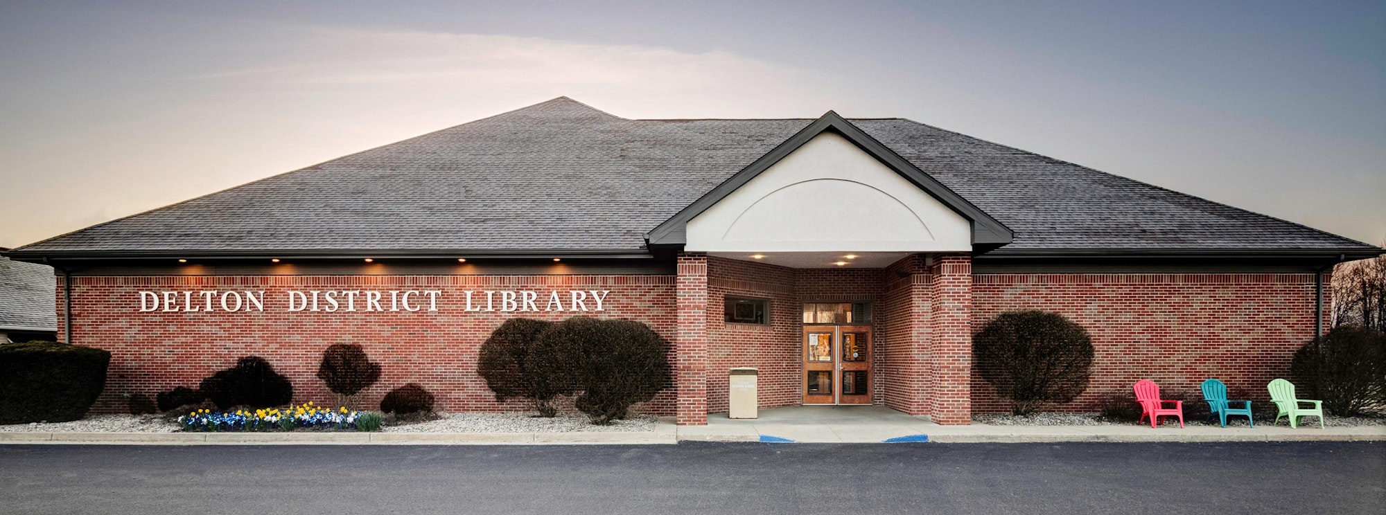 Brick library building with shrubs, colorful chairs, and visible entrance.