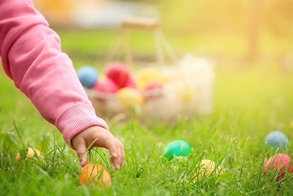 A child's hand reaches for colorful Easter eggs hidden in the grass, with a basket visible in the background.