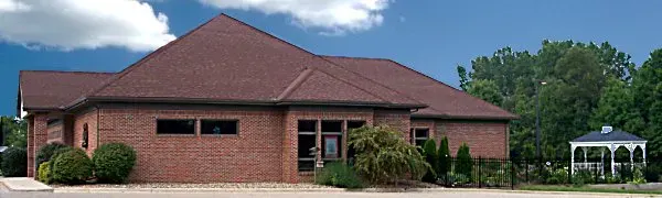 A brick building with a sloped roof, adjacent greenery, a small gazebo, and a blue sky in the background.