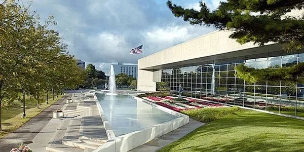 A modern building with glass walls, a fountain, American flag, trees, and landscaped gardens.