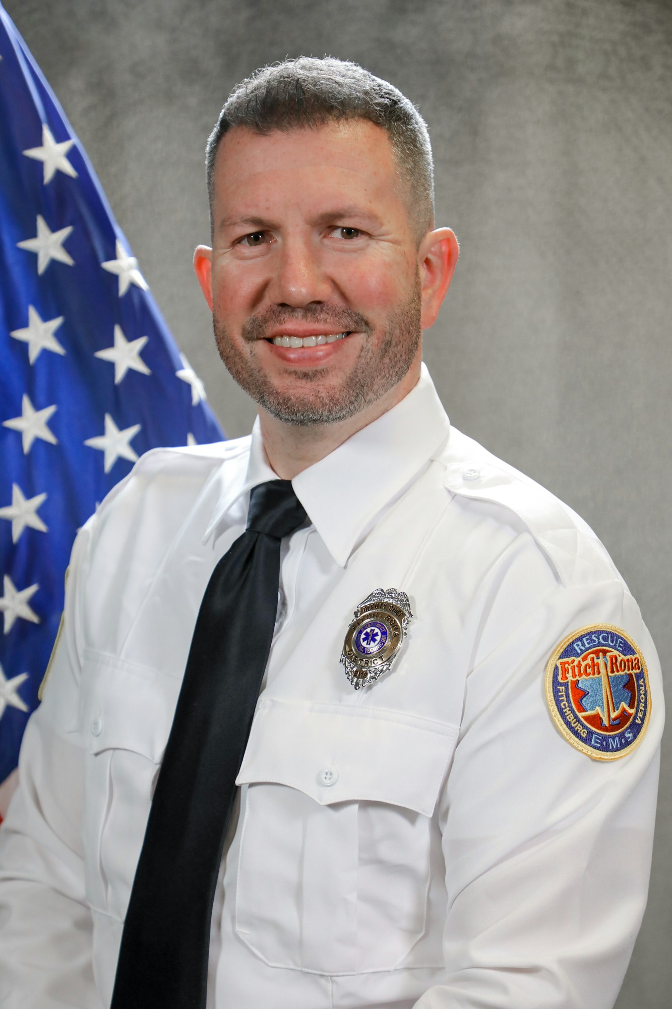 A man in uniform with a badge and "Fitch-Rona EMS" patch, smiling in front of an American flag.