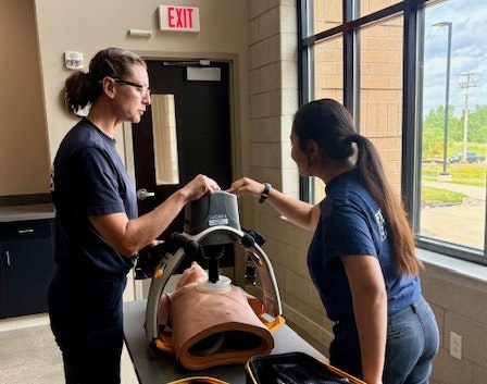 Two people practicing CPR on a medical dummy with a resuscitation device in a room with large windows.