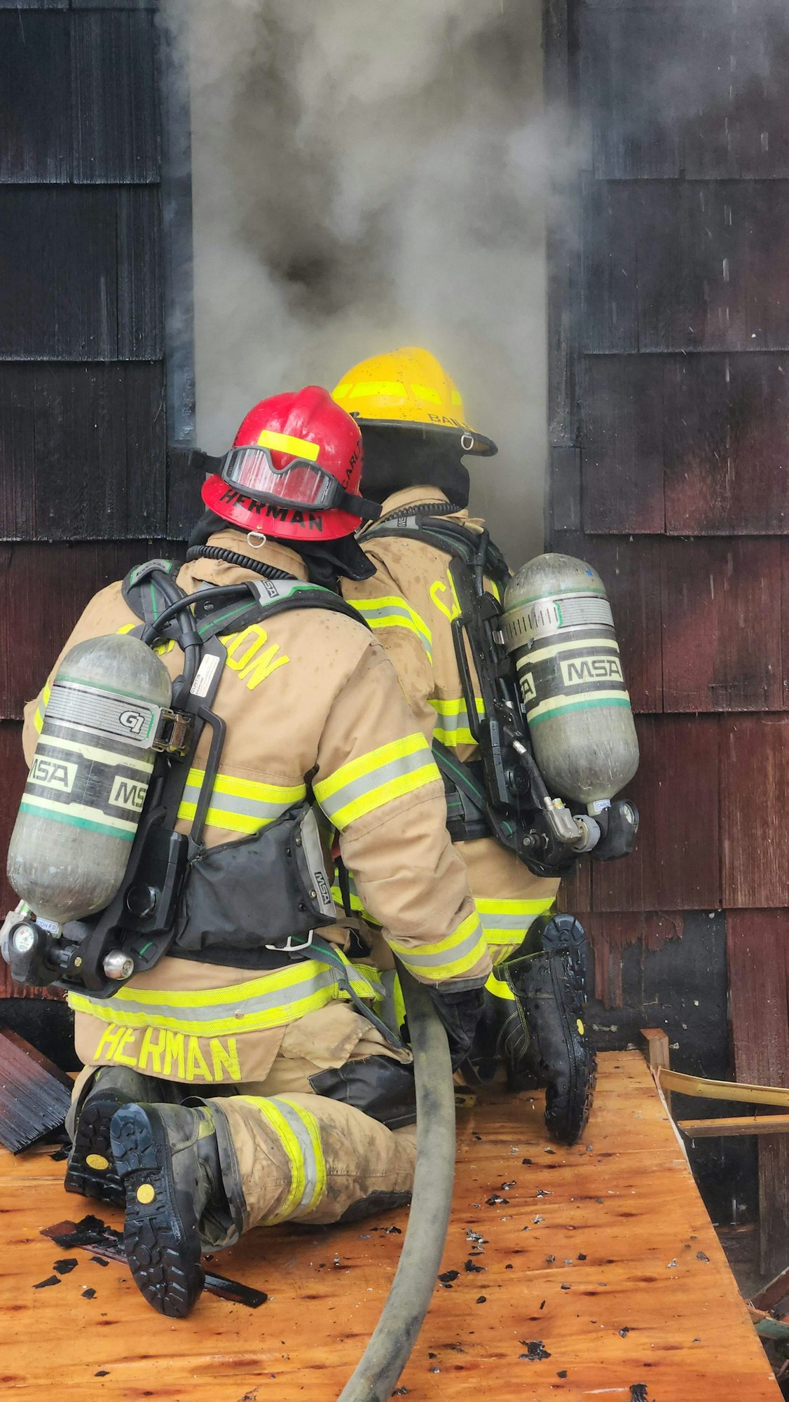 Firefighters with gear and hoses enter a smoky building through a window.