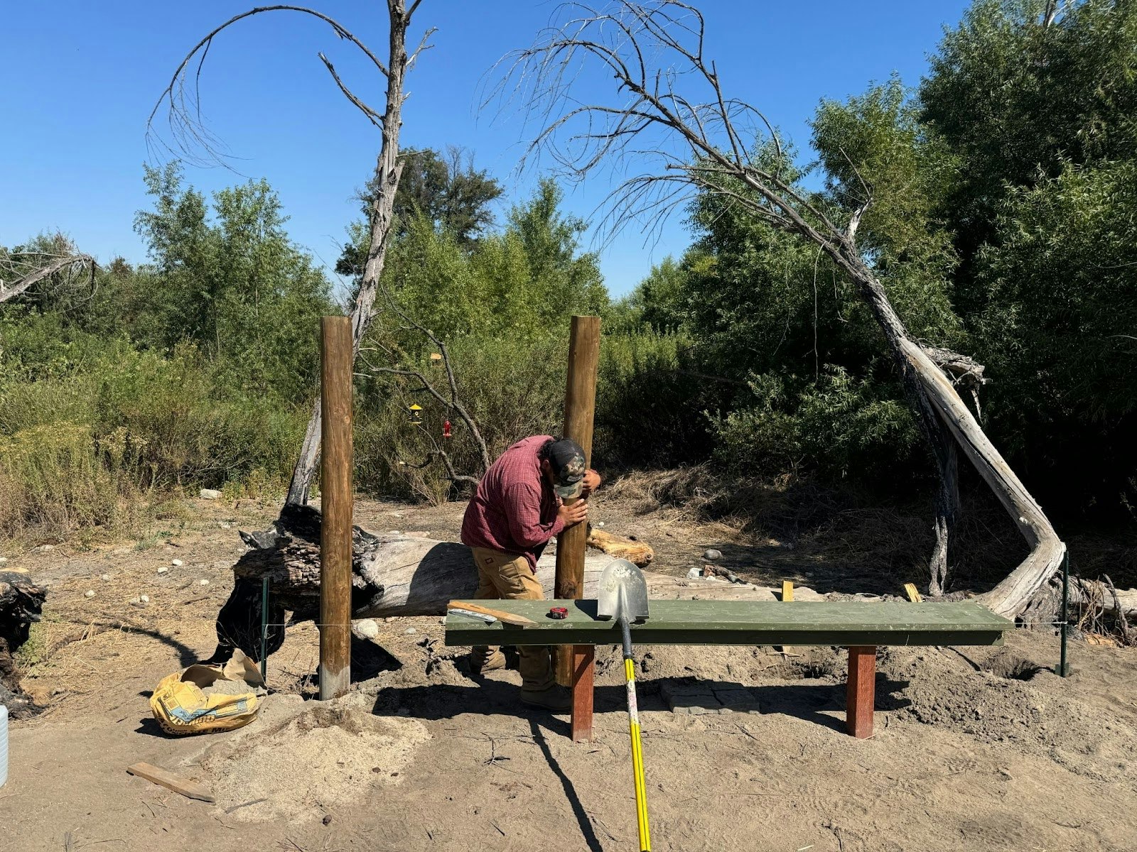 A person working outdoors assembling a wooden structure, surrounded by trees and tools.