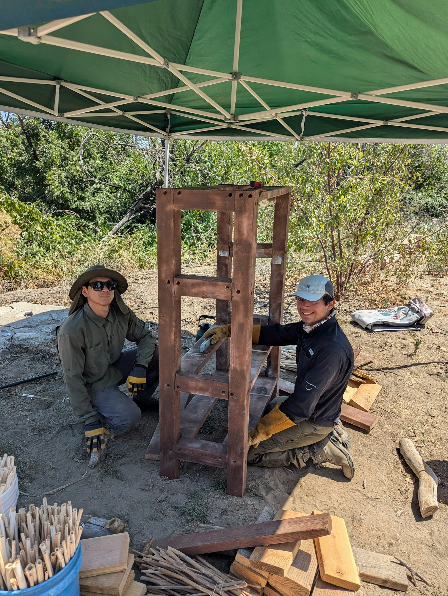 Two people under a canopy working on a wooden structure, surrounded by tools and materials outdoors.