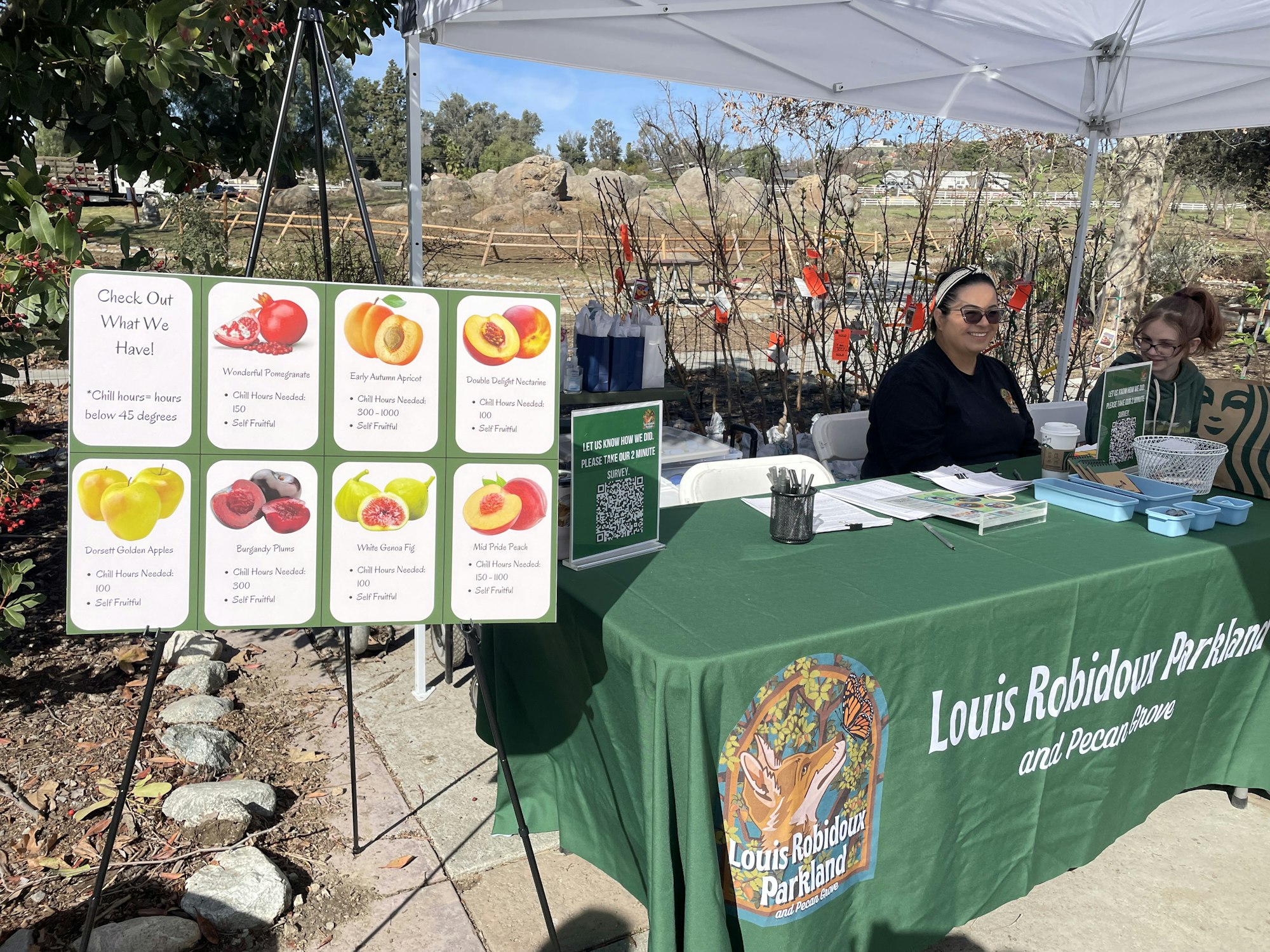 A booth with a sign listing fruit varieties and chill hours, staffed by two people under a tent with "Louis Robidoux Parkland" branding.