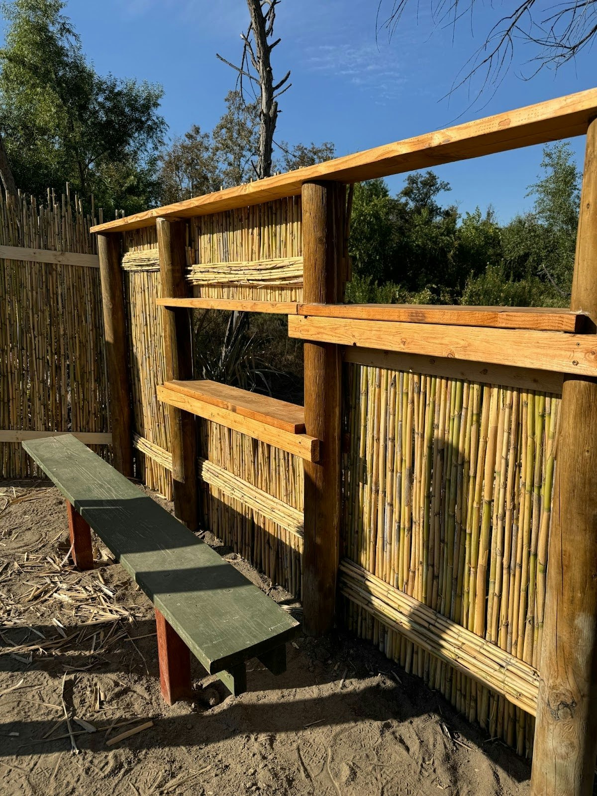 Outdoor scene with a bamboo and wood fence, a simple wooden bench, and a clear blue sky.