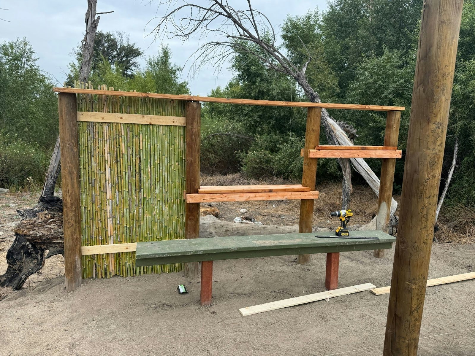 A partially constructed outdoor structure with bamboo wall, wooden shelves, green bench, and a drill on the bench, surrounded by trees.