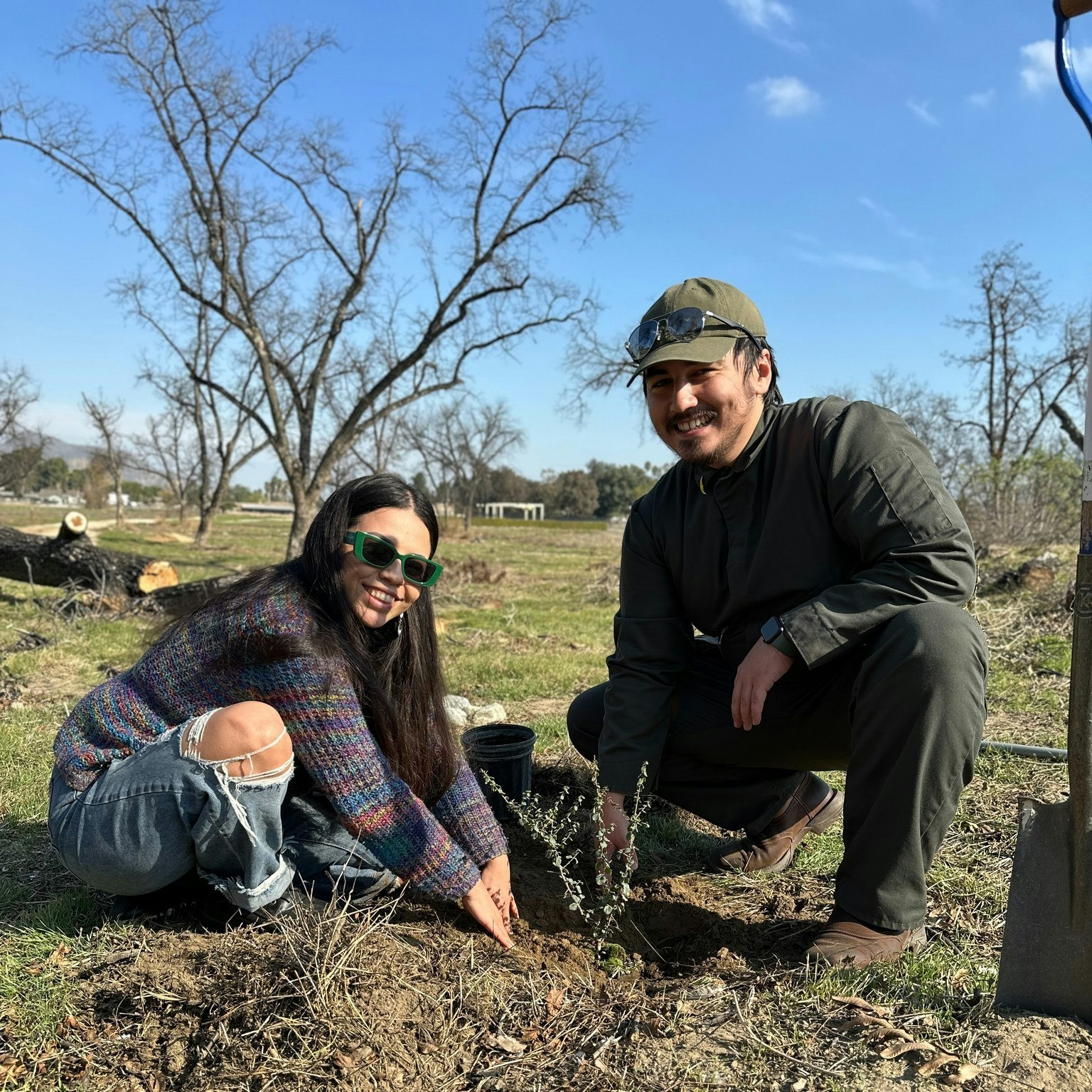Two people planting a tree outdoors, smiling, with clear skies and leafless trees in the background.