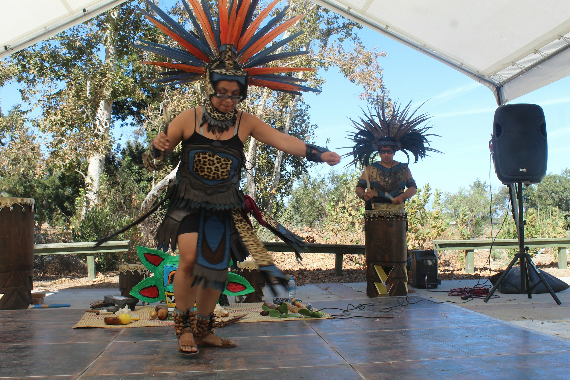 Two people in traditional attire perform a dance with drumming under a canopy outside.