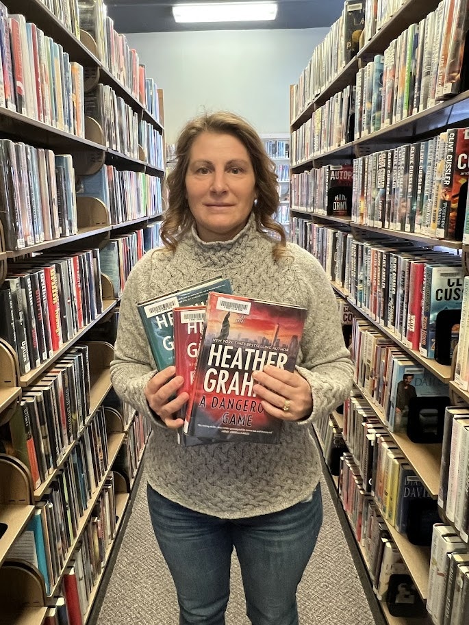A person stands in a library aisle holding three books by Heather Graham, surrounded by shelves of books.
