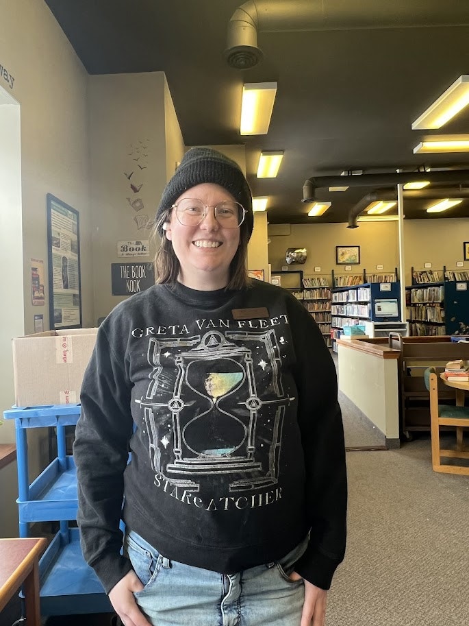 A person wearing a black sweater with "GRETA VAN FLEET STARCATCHER" stands in a library setting.