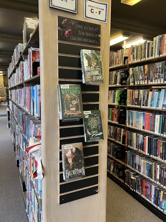 A library corner displaying books with themes of dark florals and spring, featuring titles like "Where Darkness Blooms."