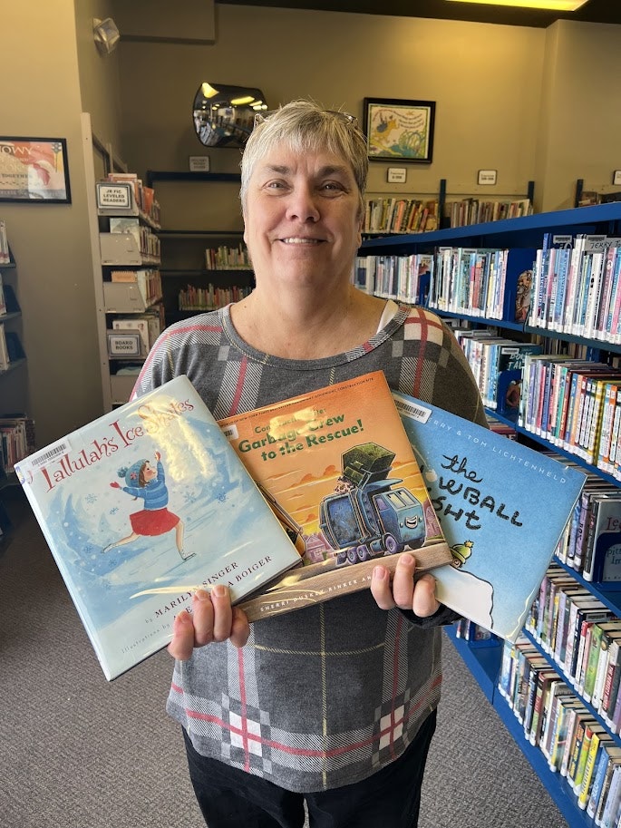 A smiling woman holds three children's books in a library, showcasing titles that encourage reading and imagination.