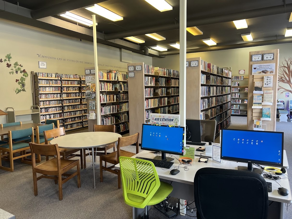 A library interior featuring computer stations, bookshelves filled with books and DVDs, and a cozy seating area.
