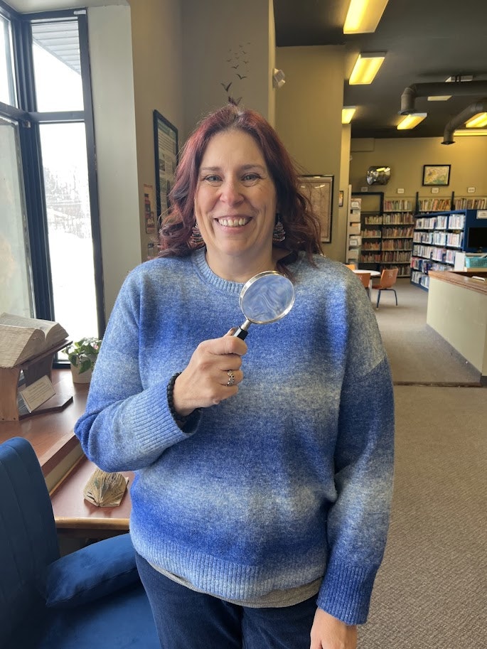 A smiling person with long hair holds a magnifying glass, standing in a library with shelves in the background.