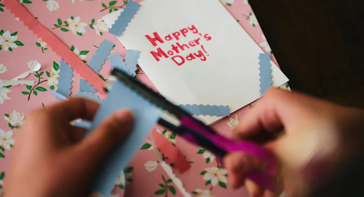A person is cutting blue paper, creating a card that says "Happy Mother’s Day!" with colorful decorations on a floral background.