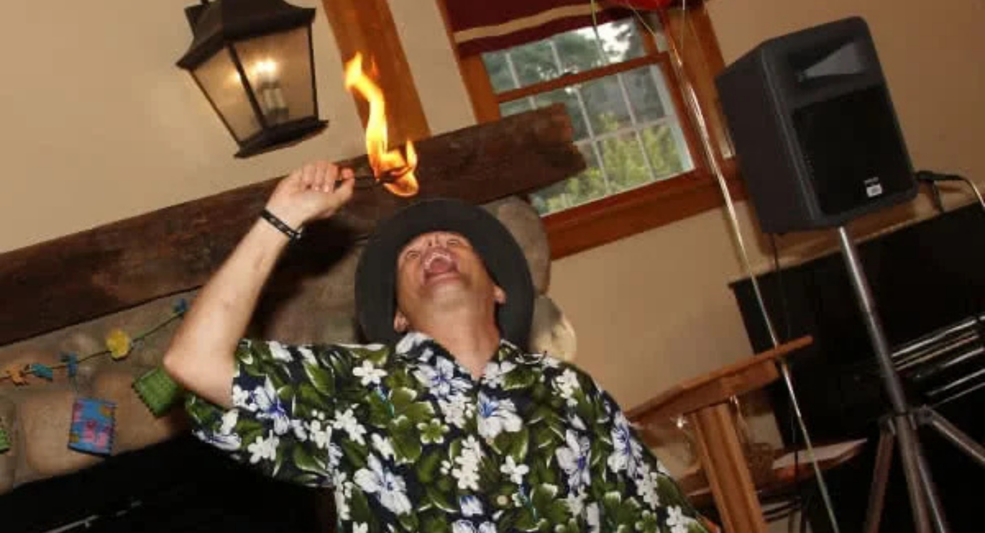 A man in a floral shirt swings a flaming object, showcasing a fire act indoors with a lively atmosphere.
