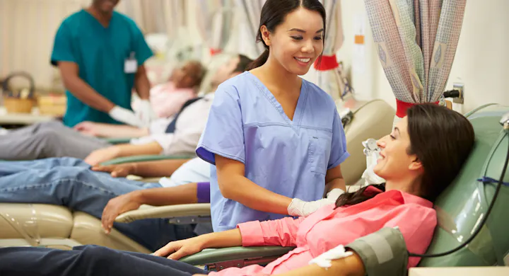 A medical professional is assisting a patient receiving treatment in a healthcare setting, with others in the background.
