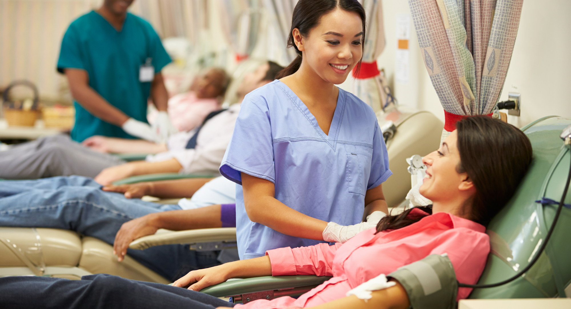 A medical professional is assisting a patient receiving treatment in a healthcare setting, with others in the background.