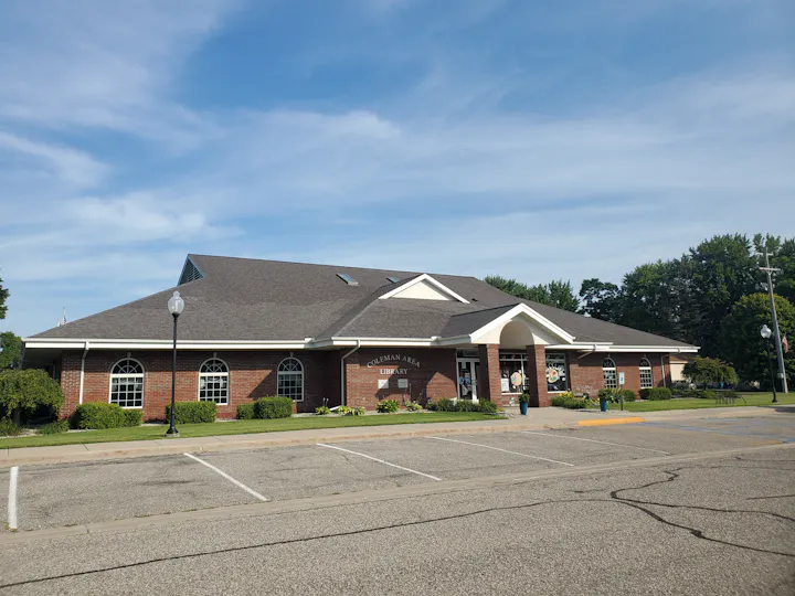 The image shows a brick library building with a peaked roof surrounded by greenery and parking spaces.