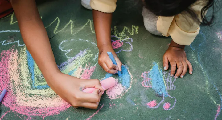 Two children are drawing colorful chalk art on a green surface, using various chalk colors to create their artwork.