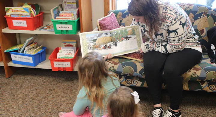 A cozy reading scene in a library, with a woman reading to two young girls on a colorful couch surrounded by books.
