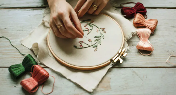 A person is embroidering a floral design with colorful threads on a fabric hoop, surrounded by spools of thread.