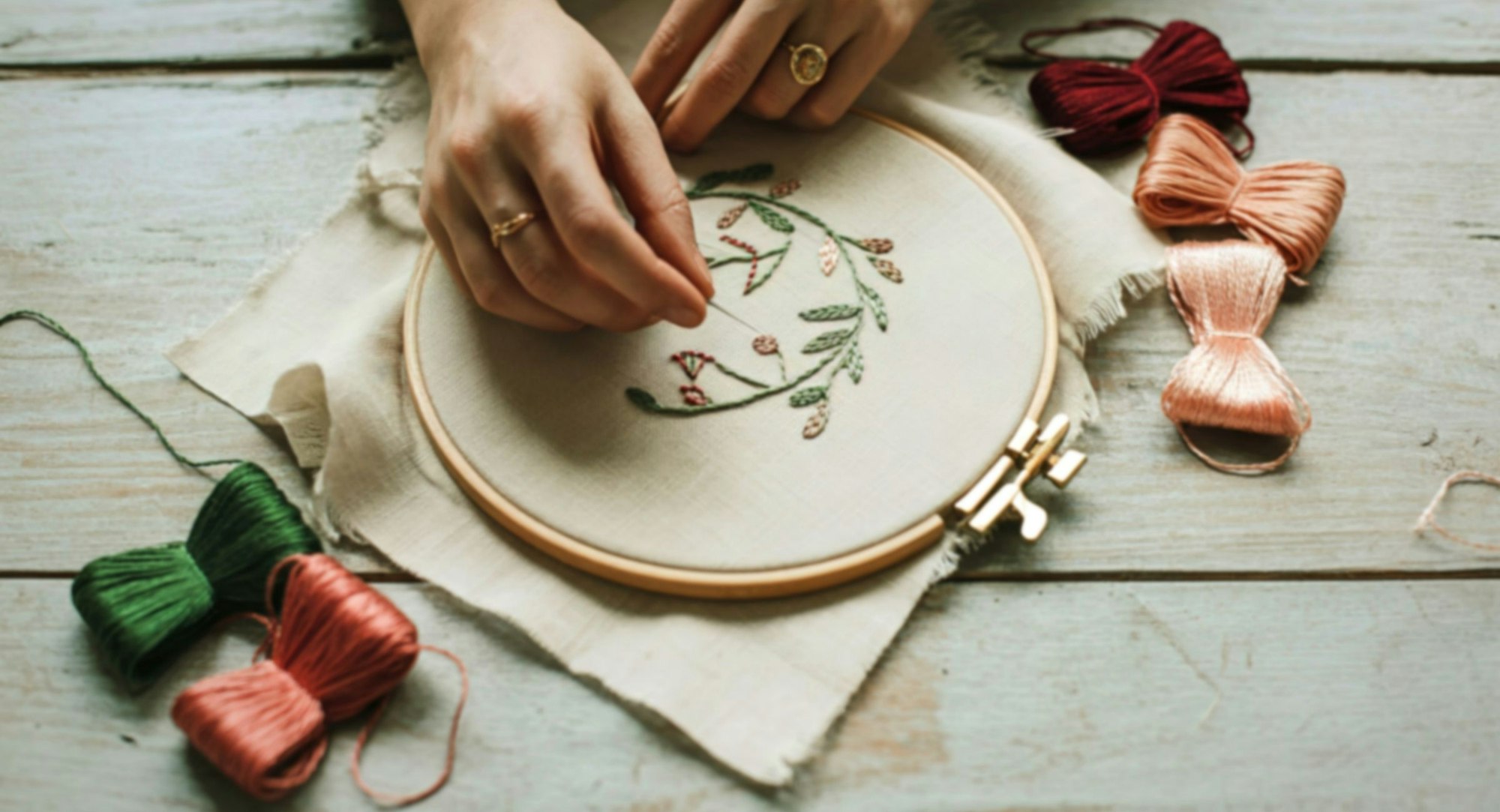 A person is embroidering a floral design with colorful threads on a fabric hoop, surrounded by spools of thread.