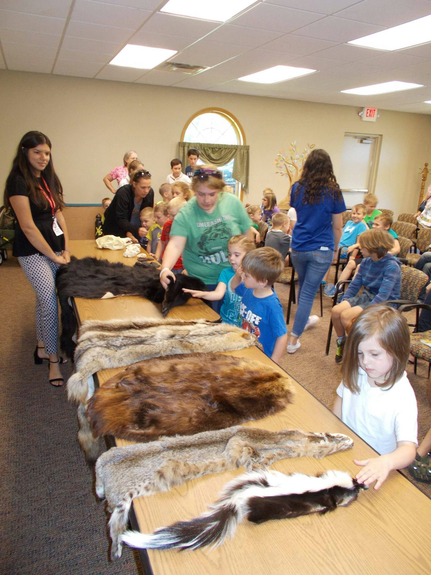 A group of children interact with various animal furs on tables during an educational session, guided by adults.