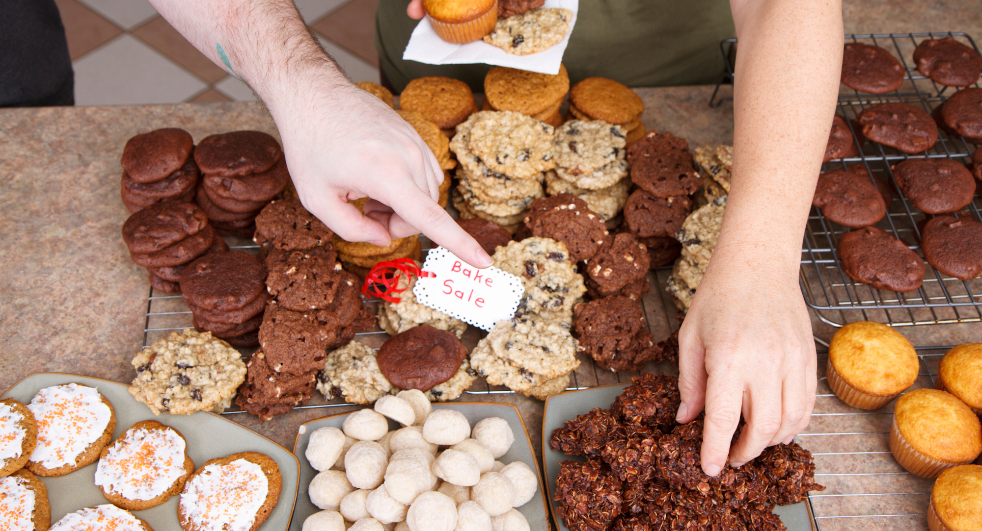 A variety of baked goods like cookies and muffins are displayed on a table, with a tag indicating a bake sale.