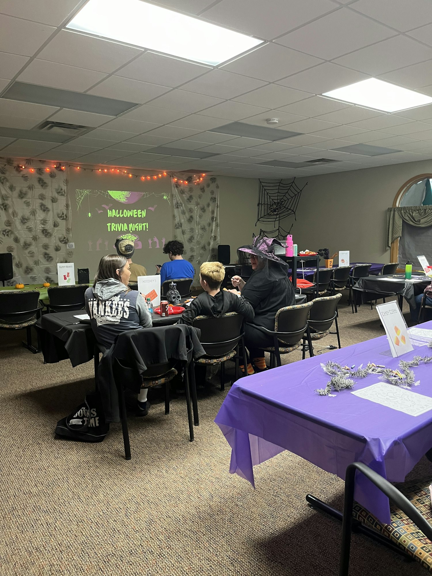The image shows a decorated room for a Halloween event, with tables, chairs, and attendees dressed in costumes.