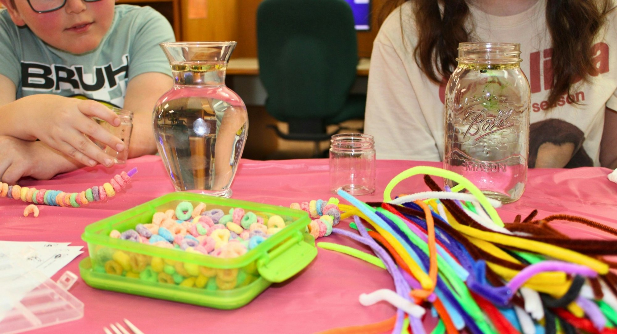 Children are engaged in an activity with jars, water, colorful cereal, and pipe cleaners on a pink tablecloth.